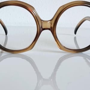 Può includere: Occhiali da vista oversize con montatura sfumata marrone e nera. Le lenti sono trasparenti e le aste si incurvano verso l'interno. Gli occhiali si riflettono su una superficie bianca, suggerendo uno stile vintage.