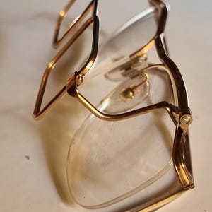 May include: Gold-coloured eyeglasses with clear lenses. The frames have a rectangular shape and are folded. The temples are thin and curve around the ears. The glasses are resting on a white surface.