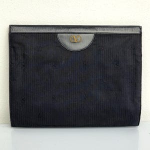 May include: A black clutch with a vertical ribbed texture. The top features a dark leather panel with a gold-colored logo. The clutch has a rectangular shape and appears to be made of a fabric material.