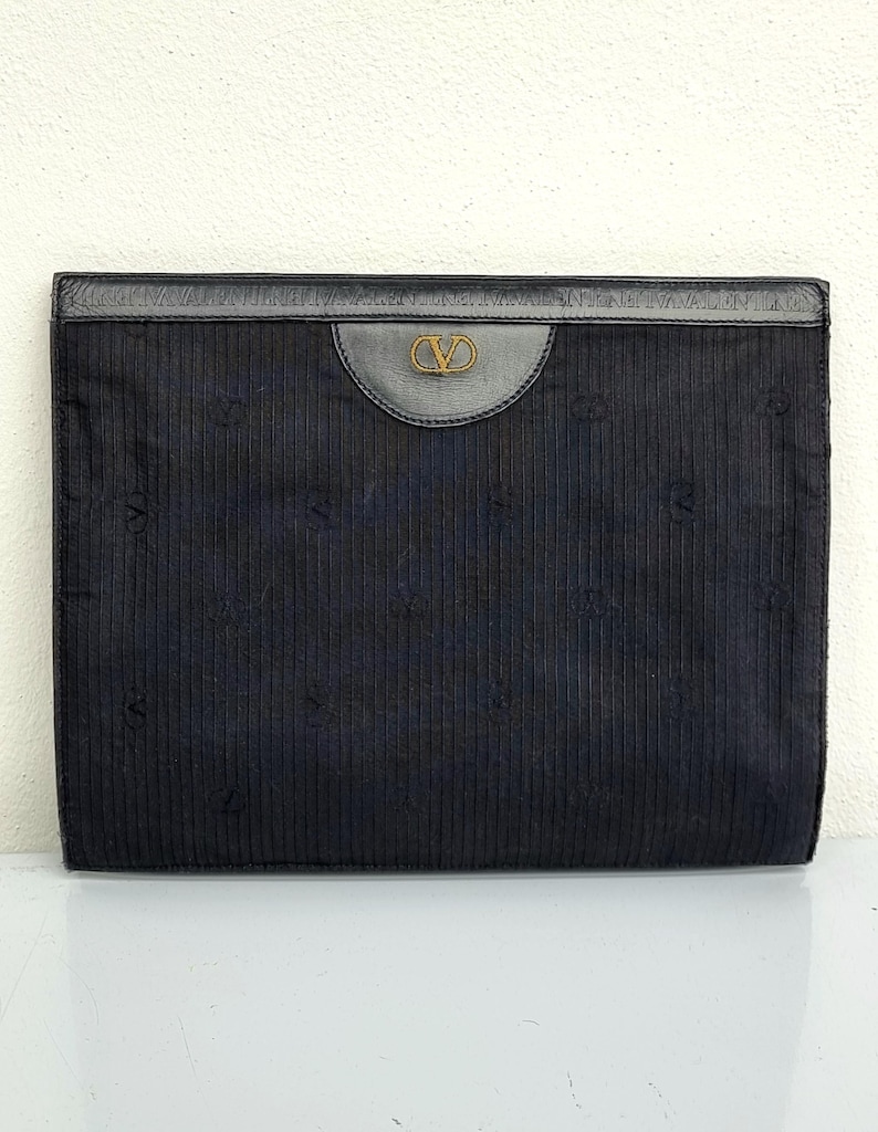 May include: A black clutch with a vertical ribbed texture. The top features a dark leather panel with a gold-colored logo. The clutch has a rectangular shape and appears to be made of a fabric material.