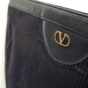 May include: Close-up of a navy blue clutch bag. The bag features a textured fabric with a leather flap. The flap has a gold embroidered "V" logo and the brand name embossed on the leather.