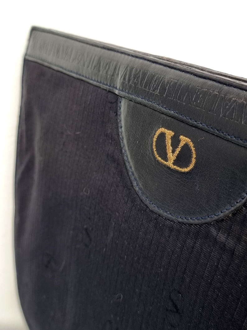 May include: Close-up of a navy blue clutch bag. The bag features a textured fabric with a leather flap. The flap has a gold embroidered "V" logo and the brand name embossed on the leather.