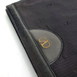 May include: A navy blue clutch with a leather handle and a circular emblem. The emblem features a gold embroidered logo. The clutch has a textured fabric with a repeating pattern of the logo.