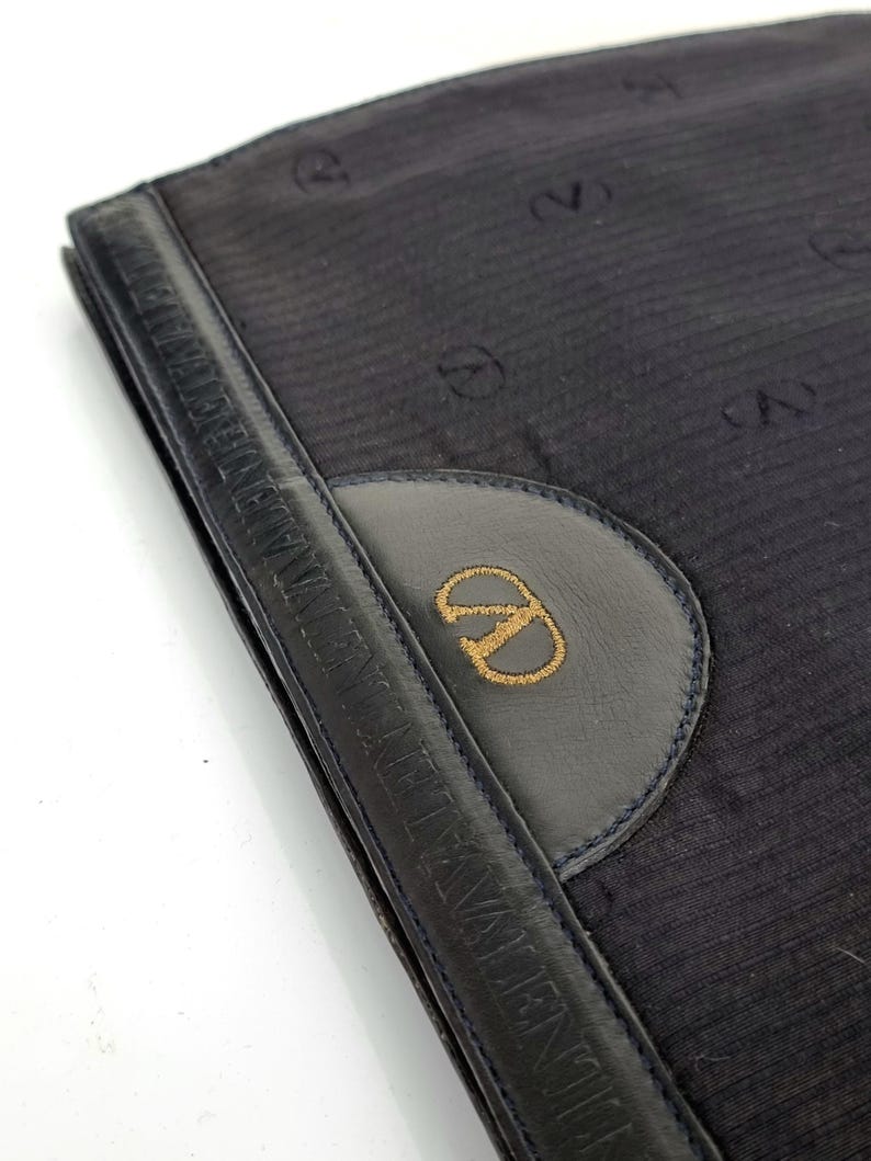 May include: A navy blue clutch with a leather handle and a circular emblem. The emblem features a gold embroidered logo. The clutch has a textured fabric with a repeating pattern of the logo.