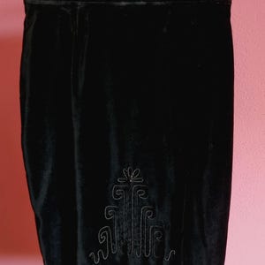May include: A black velvet skirt with an embroidered design near the hem. The skirt is hanging on a black hanger against a pink background. The embroidered design is a stylized floral pattern.