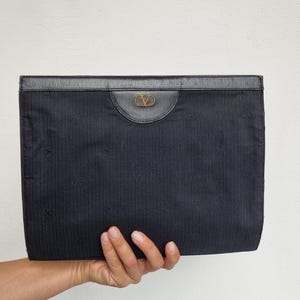 May include: A navy blue clutch with a textured fabric and a leather top. The clutch features a gold-colored logo on the front. The bag is held by a hand against a white background.