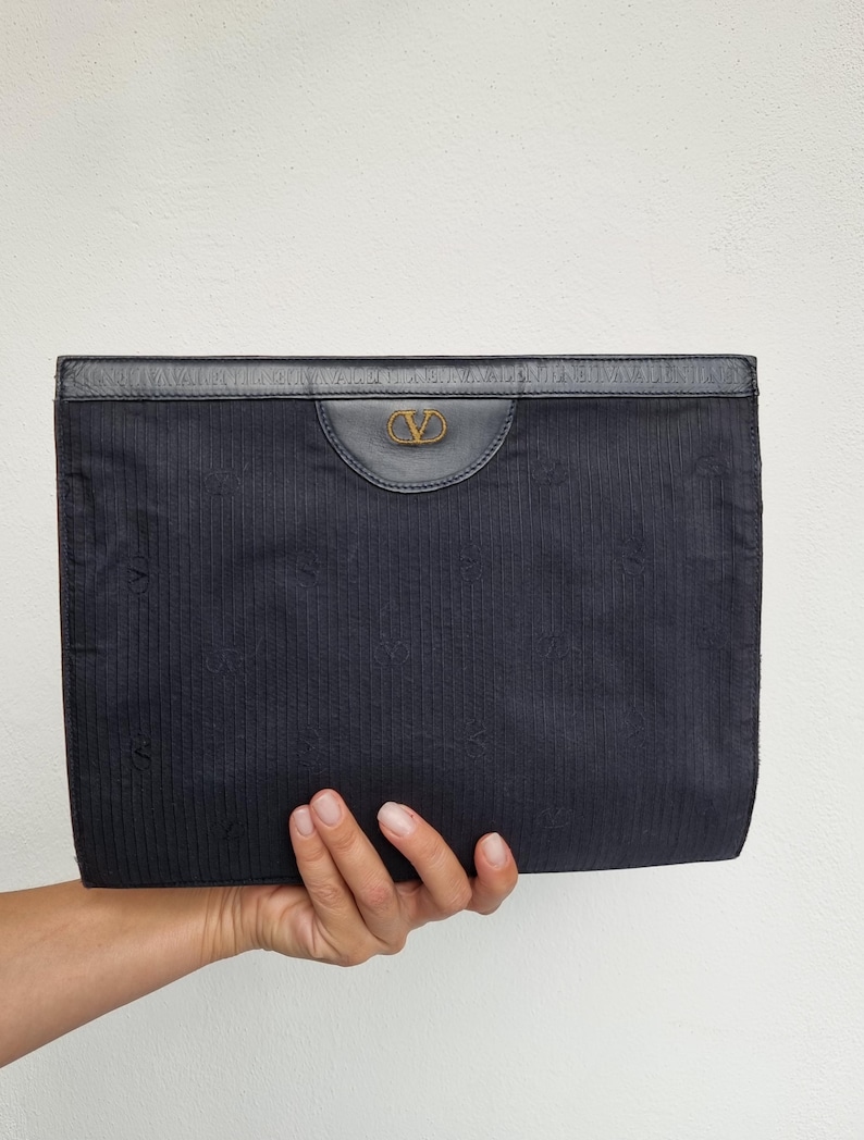 May include: A navy blue clutch with a textured fabric and a leather top. The clutch features a gold-colored logo on the front. The bag is held by a hand against a white background.