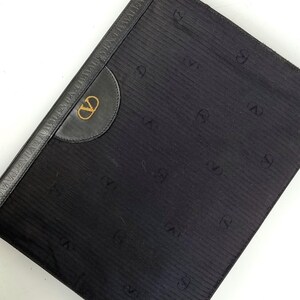 May include: Black clutch with a textured, vertically striped pattern. Features a leather accent with a gold-colored logo and a leather border with embossed text. The design includes repeating "V" symbols.