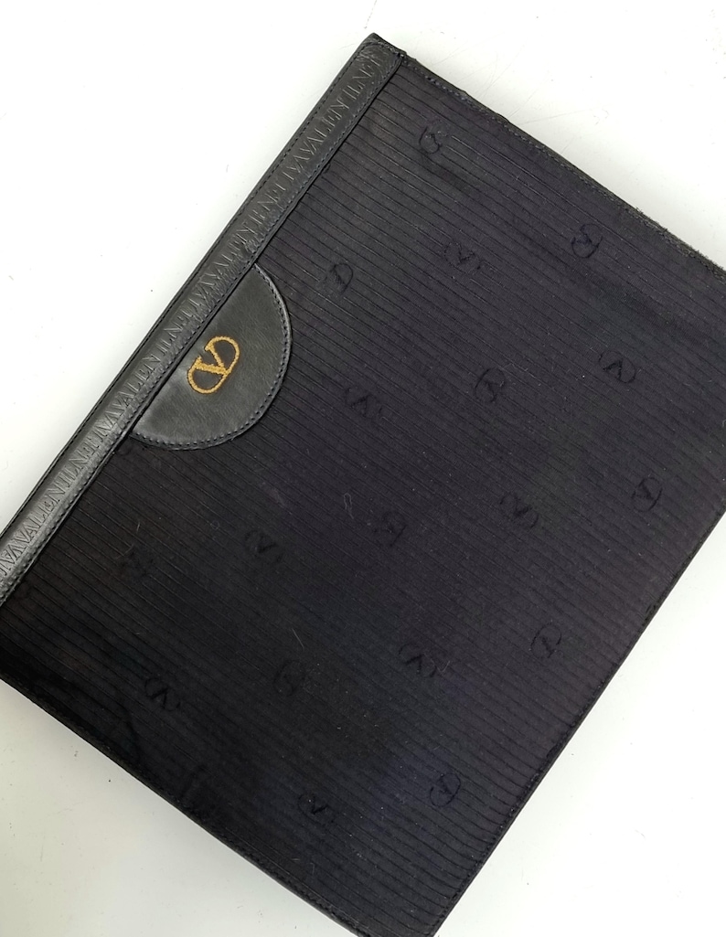 May include: Black clutch with a textured, vertically striped pattern. Features a leather accent with a gold-colored logo and a leather border with embossed text. The design includes repeating "V" symbols.