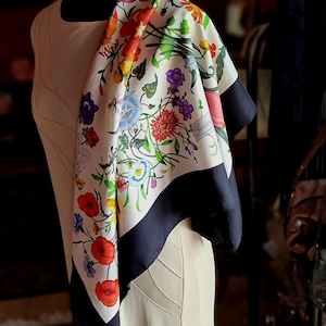 May include: A square silk scarf featuring a white background with a vibrant floral print of red, orange, yellow, blue, and purple flowers and green foliage. The scarf has a wide black border and the Gucci brand name printed in cursive script.
