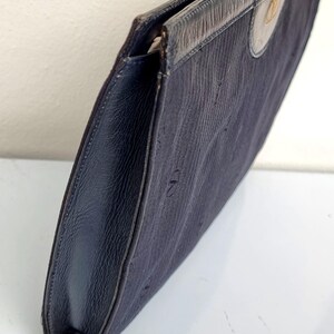 May include: A navy blue clutch with a textured fabric body and leather trim. The clutch features a flap closure and a gold-colored emblem on the front. The edges are dark blue leather.