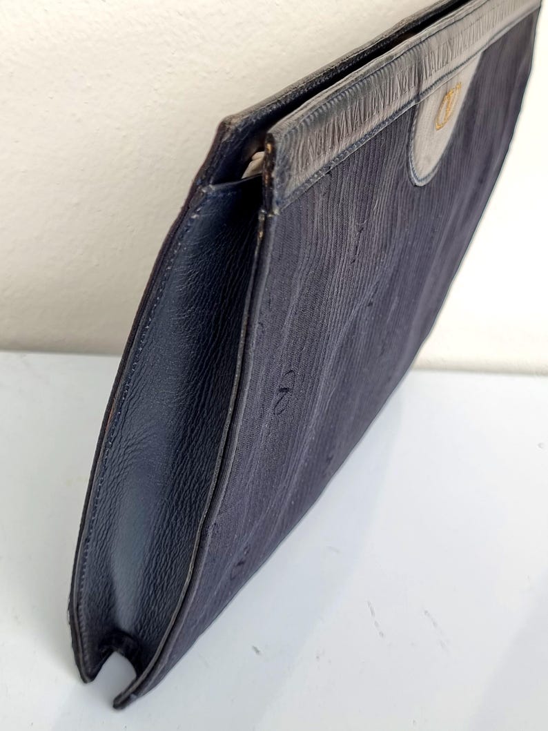 May include: A navy blue clutch with a textured fabric body and leather trim. The clutch features a flap closure and a gold-colored emblem on the front. The edges are dark blue leather.