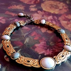 Può includere: Una collana dorata con motivi a nodo celtico e pietre preziose ovali bianche e grigie. La collana ha una chiusura a catena ed è esposta su uno sfondo floreale. Il gioiello è probabilmente vintage o ispirato all'arte celtica.