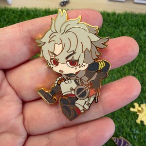 May include: A gold-colored enamel pin featuring a stylized anime character with gray hair, red eyes, and a determined expression. The character is dressed in a white and black outfit with gold accents. The pin is held in a hand.