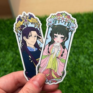 May include: Two stickers featuring anime-style illustrations of characters in traditional Asian attire. One sticker depicts a person with long dark hair and a blue and black outfit. The other shows a person in a pink and yellow outfit.