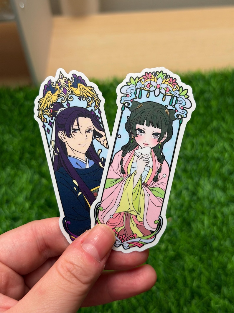 May include: Two stickers featuring anime-style illustrations of characters in traditional Asian attire. One sticker depicts a person with long dark hair and a blue and black outfit. The other shows a person in a pink and yellow outfit.