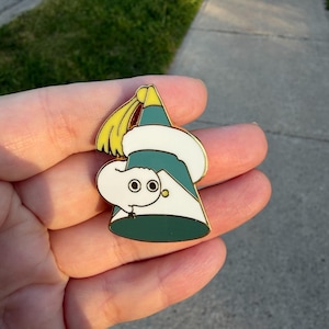 May include: A metal enamel pin shaped like a teal party hat with a white fluffy trim and a yellow tassel at the top. A small white worm-like creature with two large black eyes is wrapped around the front of the hat, peering out from the side.