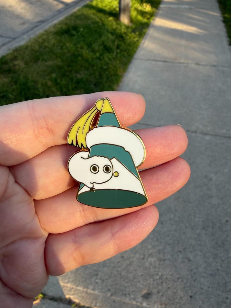 May include: A metal enamel pin shaped like a teal party hat with a white fluffy trim and a yellow tassel at the top. A small white worm-like creature with two large black eyes is wrapped around the front of the hat, peering out from the side.