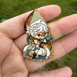 May include: A gold-toned metal enamel pin featuring a stylized character with white hair, wearing a pointed hat and white robes, holding an open book with a gold star emblem, surrounded by blue wave-like patterns and gold outlines.