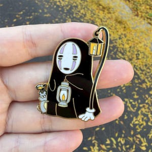 K&ouml;nnte beinhalten: Ein schwarzer Emaille-Pin mit einer Figur aus Spirited Away, die eine Laterne und eine kleine Kreatur h&auml;lt. Der Pin hat goldfarbene Akzente und ein wei&szlig;es Gesicht mit lila Markierungen. Die Figur tr&auml;gt einen schwarzen Umhang.