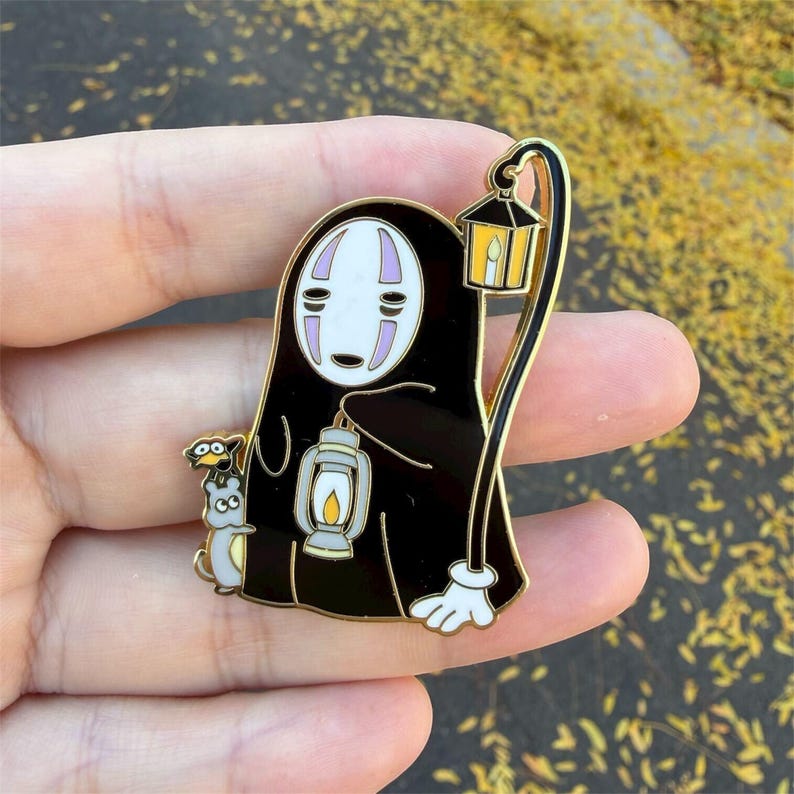 K&ouml;nnte beinhalten: Ein schwarzer Emaille-Pin mit einer Figur aus Spirited Away, die eine Laterne und eine kleine Kreatur h&auml;lt. Der Pin hat goldfarbene Akzente und ein wei&szlig;es Gesicht mit lila Markierungen. Die Figur tr&auml;gt einen schwarzen Umhang.