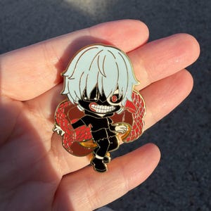 K&ouml;nnte beinhalten: Ein farbenfroher Emaille-Pin mit einem Charakter im Chibi-Stil mit wei&szlig;en Haaren, einer schwarzen Maske und einem roten Auge. Die Figur tr&auml;gt ein schwarzes Outfit und ist von einem roten und braunen Design umgeben. Der Pin ist wahrscheinlich ein Sammlerst&uuml;ck.
