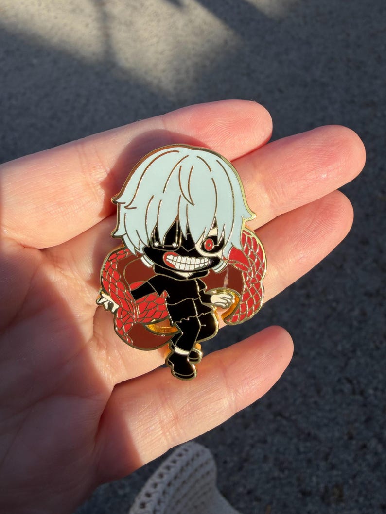 K&ouml;nnte beinhalten: Ein farbenfroher Emaille-Pin mit einem Charakter im Chibi-Stil mit wei&szlig;en Haaren, einer schwarzen Maske und einem roten Auge. Die Figur tr&auml;gt ein schwarzes Outfit und ist von einem roten und braunen Design umgeben. Der Pin ist wahrscheinlich ein Sammlerst&uuml;ck.