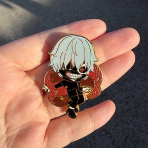 K&ouml;nnte beinhalten: Ein goldfarbener Emaille-Pin mit einem Charakter im Chibi-Stil mit hellblauen Haaren, einer schwarzen Maske und einem roten Auge. Die Figur tr&auml;gt ein schwarzes Outfit und ist von einem rot-braunen Design umgeben. Der Pin ist wahrscheinlich ein Sammlerst&uuml;ck.
