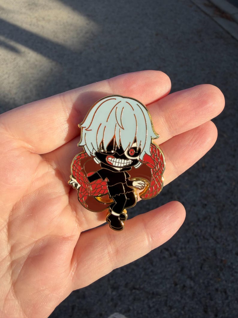 K&ouml;nnte beinhalten: Ein goldfarbener Emaille-Pin mit einem Charakter im Chibi-Stil mit hellblauen Haaren, einer schwarzen Maske und einem roten Auge. Die Figur tr&auml;gt ein schwarzes Outfit und ist von einem rot-braunen Design umgeben. Der Pin ist wahrscheinlich ein Sammlerst&uuml;ck.