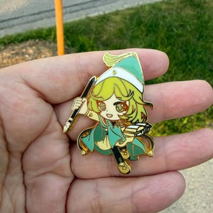 May include: A gold-toned metal enamel pin featuring a stylized cartoon character with light green hair, wearing a matching pointed hat and cape. The character holds a black quill pen in one hand and an open book in the other, with gold outlines.