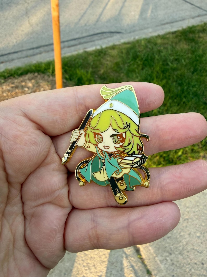 May include: A gold-toned metal enamel pin featuring a stylized cartoon character with light green hair, wearing a matching pointed hat and cape. The character holds a black quill pen in one hand and an open book in the other, with gold outlines.