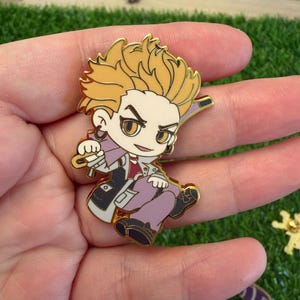 May include: A gold-colored enamel pin featuring a cartoon character with spiky yellow hair, wearing a purple outfit with a red shirt and black shoes. The pin has a gold border and is held in a hand.