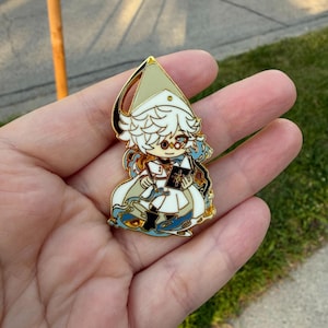 May include: A gold-toned metal enamel pin featuring a stylized character with white hair, wearing a pointed hat and white robes, holding a black book with a gold star emblem, surrounded by swirling blue and gold decorative elements.