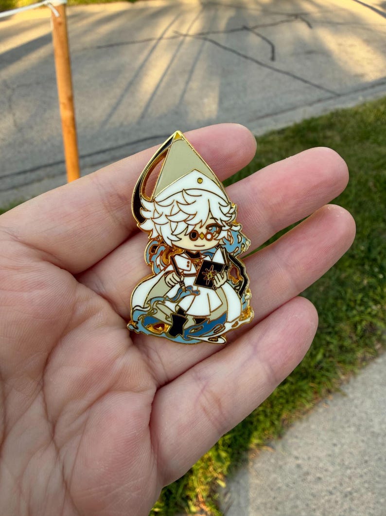 May include: A gold-toned metal enamel pin featuring a stylized character with white hair, wearing a pointed hat and white robes, holding a black book with a gold star emblem, surrounded by swirling blue and gold decorative elements.