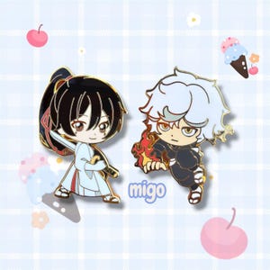 May include: Two enamel pins featuring anime-style characters. One pin depicts a character with black hair and a sword, wearing a white outfit. The other pin shows a character with white hair and flames, wearing a black outfit. The word "migo" is printed on the image.
