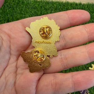 May include: Two gold-colored enamel pins with butterfly-shaped designs. Each pin has a gold clasp on the back for secure attachment. The pins are displayed against a backdrop of green artificial grass.