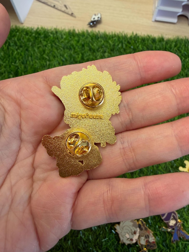 May include: Two gold-colored enamel pins with butterfly-shaped designs. Each pin has a gold clasp on the back for secure attachment. The pins are displayed against a backdrop of green artificial grass.