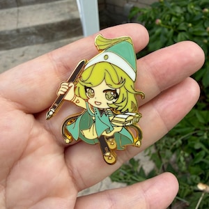 May include: A gold-toned metal enamel pin featuring a stylized anime-inspired character with light green hair, wearing a matching green hat and cape. The character holds a dark brown fountain pen in one hand and an open book in the other.