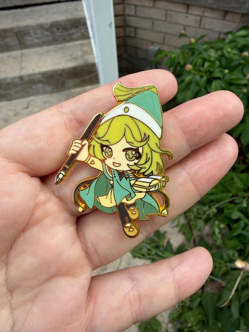 May include: A gold-toned metal enamel pin featuring a stylized anime-inspired character with light green hair, wearing a matching green hat and cape. The character holds a dark brown fountain pen in one hand and an open book in the other.