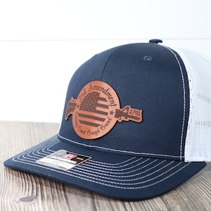 May include: A navy blue and white trucker hat with a brown leather patch that says "2nd Amendment" with an American flag and a gun graphic. The patch also says "1791" and "God Given Right Guns".