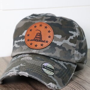 May include: A camouflage baseball cap with a distressed look. The cap features a brown leather patch with a snake and stars, and the text "KETHOS Vintage Since 2001".