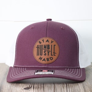 May include: A burgundy and white trucker hat with a brown leather patch that says "Stay Humble Hustle Hard".