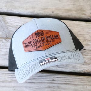 May include: A light blue and black trucker hat with a brown leather patch. The patch features the text "BLUE COLLAR DOLLAR" and the phrase "I WASN'T BORN WITH IT; I WORKED FOR IT." An American flag is also on the patch.