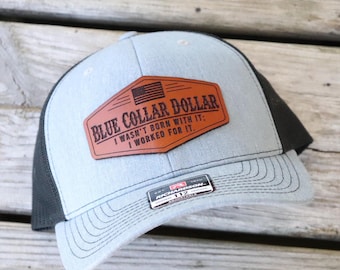 May include: A light blue and black trucker hat with a brown leather patch. The patch features the text "BLUE COLLAR DOLLAR" and the phrase "I WASN'T BORN WITH IT; I WORKED FOR IT." An American flag is also on the patch.