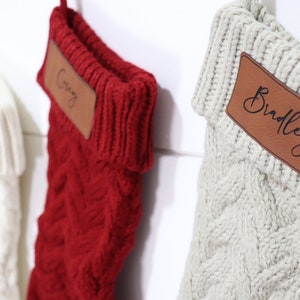May include: Close-up of three cable-knit Christmas stockings in white, red, and off-white. Each stocking has a brown leather patch with a name on it. The names are "Casey" and "Bradley".