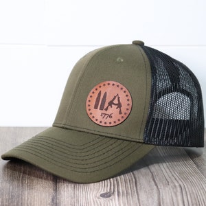 May include: A green and black trucker hat with a brown leather patch featuring a silhouette of three rifles and the text "1776". The patch is surrounded by a circle of stars.