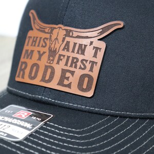 May include: A black and gray trucker hat with a brown leather patch that says "This Ain't My First Rodeo" with a skull and horns design. The hat is adjustable and has a Richardson logo.