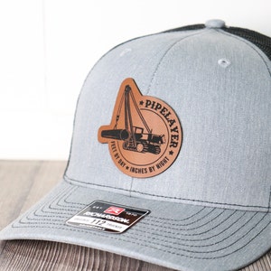 May include: A gray and black trucker hat with a brown leather patch featuring a silhouette of a pipe laying machine and the text "PIPELAYER * FEET BY DAY * INCHES BY NIGHT *". The hat has a Richardson 112 Trucker label.