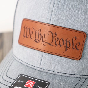 May include: A gray baseball cap with a brown leather patch that says "We the People" in a cursive font.