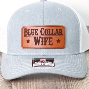 May include: A gray and white trucker hat with a brown leather patch that says "BLUE COLLAR WIFE" with stars on either side. The hat is adjustable and has a Richardson 112 logo.